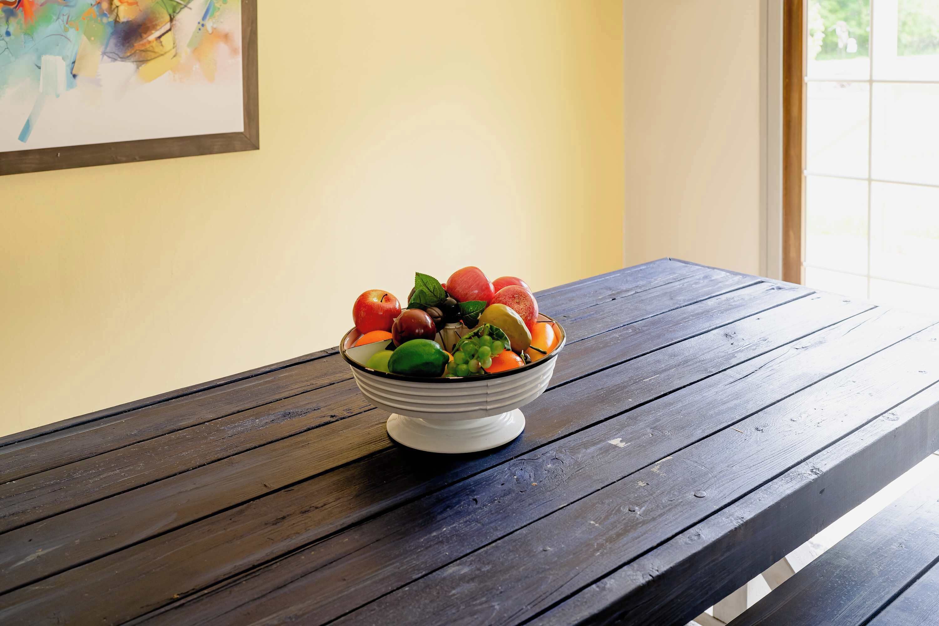 A bowl of fruit on a wooden table with a painting on the wall.