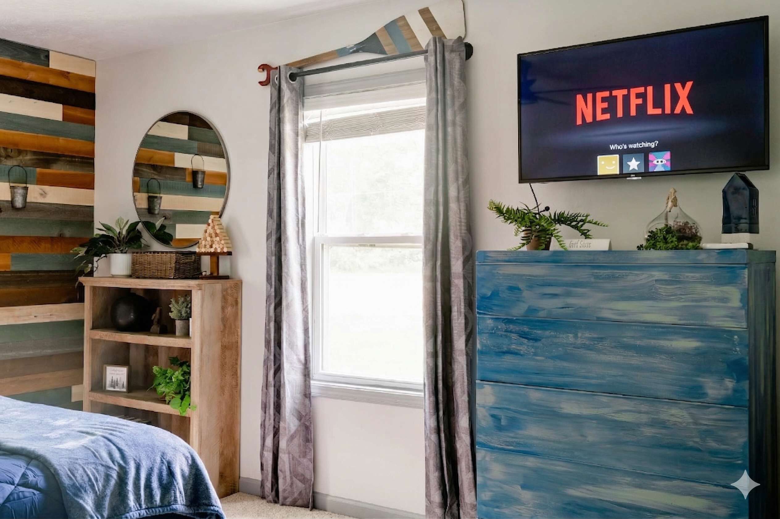 A bedroom with a bed, a TV mounted on the wall displaying 'NETFLIX,' a window with curtains, a wooden shelf with plants, and a blue dresser.