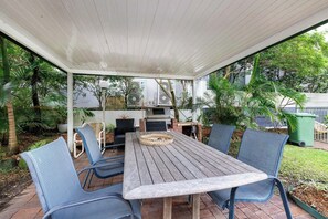 An outdoor patio with a wooden table and chairs, a grill, and air conditioning units. - Coastal 1BDR Flat with Pool, Burleigh Heads (Burleigh Heads)