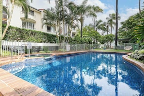 Une piscine entourée d’une clôture, de palmiers et d’un bâtiment doté de balcons.