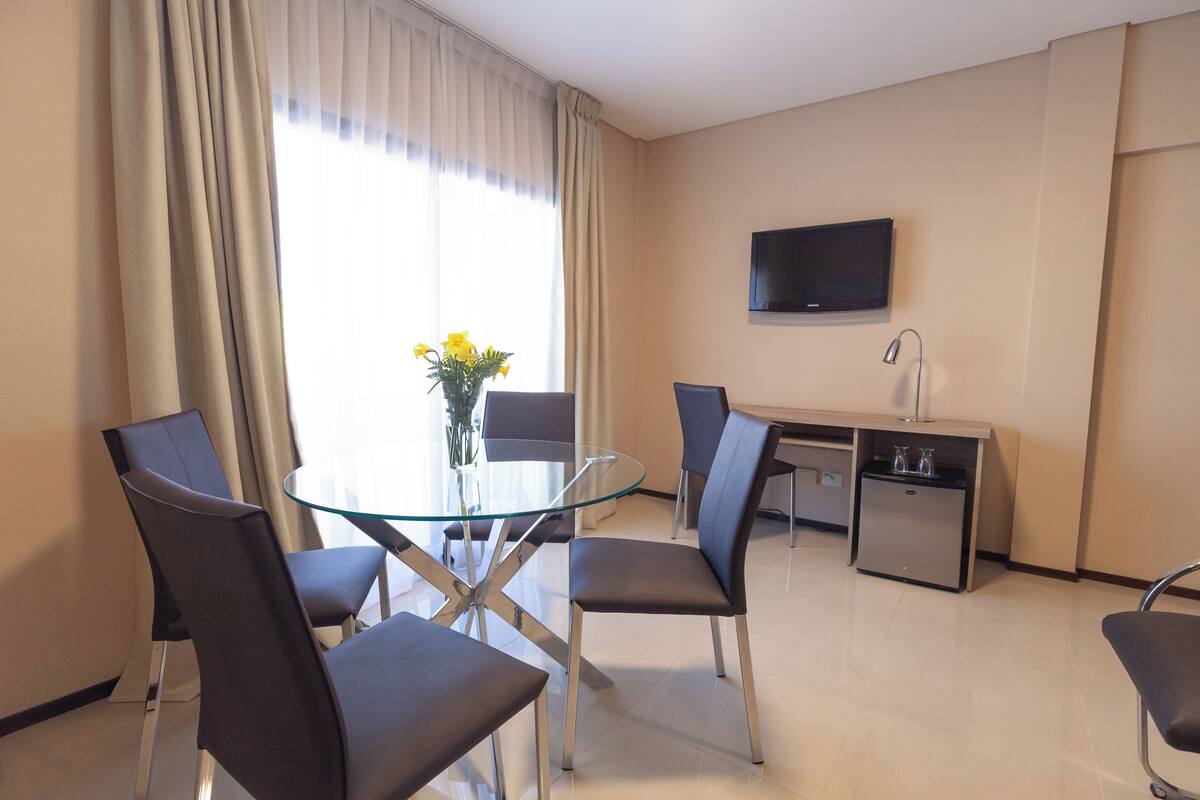 Habitación de hotel moderna con una mesa de comedor de vidrio, cuatro sillas, un escritorio pequeño con un televisor y un mini bar.