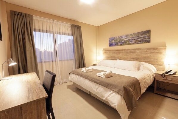 A hotel room with a bed, a desk, a chair, a nightstand with a phone, and a painting on the wall. - Telepostal Congreso (Buenos Aires)