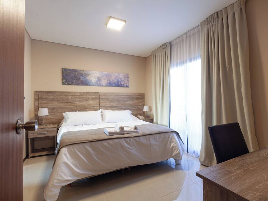 Quarto de hotel com cama grande, mesas de cabeceira com luminárias, cabeceira de madeira, um quadro na parede e vista para o exterior através de cortinas.
