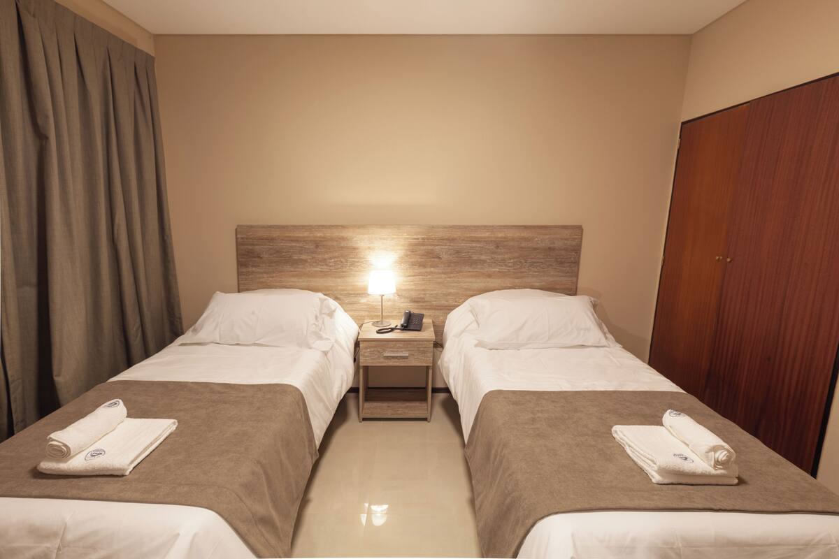 Habitación de hotel con dos camas, cabecera de madera, mesita de noche con lámpara y armario.