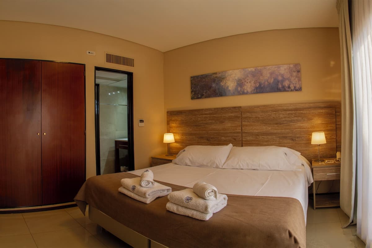Habitación de hotel con una cama grande, dos mesitas de noche con lámparas, un cabecero de madera, un cuadro en la pared y una puerta.