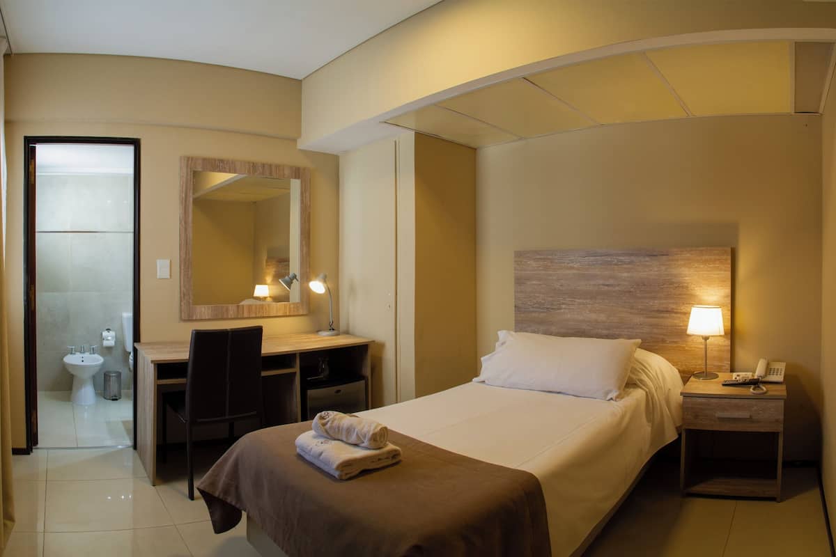 Habitación de hotel con cama, escritorio, silla y baño visible a través de una puerta abierta.