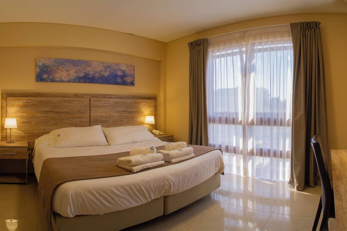 Habitación de hotel con una cama grande, dos lámparas de noche, un cuadro en la pared y vistas a través de cortinas translúcidas.
