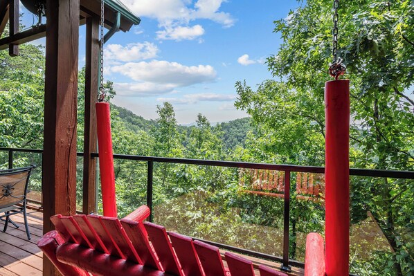 A wooden deck with red chairs and a table, overlooking a lush green forest and a river.