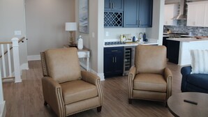 A modern living room with two leather armchairs, a coffee table, and a view into a kitchen with dark blue cabinets and a white countertop. - BRAND NEW HOME, PRIVATE POOL, partial ocean view  By Williamson Realty (Ocean Isle Beach)