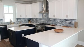 A modern kitchen with white cabinets, a central island, a stainless steel range hood, and a tiled backsplash. - BRAND NEW HOME, PRIVATE POOL, partial ocean view  By Williamson Realty (Ocean Isle Beach)