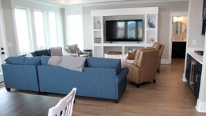 A modern living room with a blue sofa, a tan armchair, and a flat-screen TV on a white entertainment unit. - BRAND NEW HOME, PRIVATE POOL, partial ocean view  By Williamson Realty (Ocean Isle Beach)