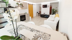 A modern kitchen with a built-in oven, a dining area with a patterned tablecloth, and a living room with a white sofa and a TV mounted on the wall.