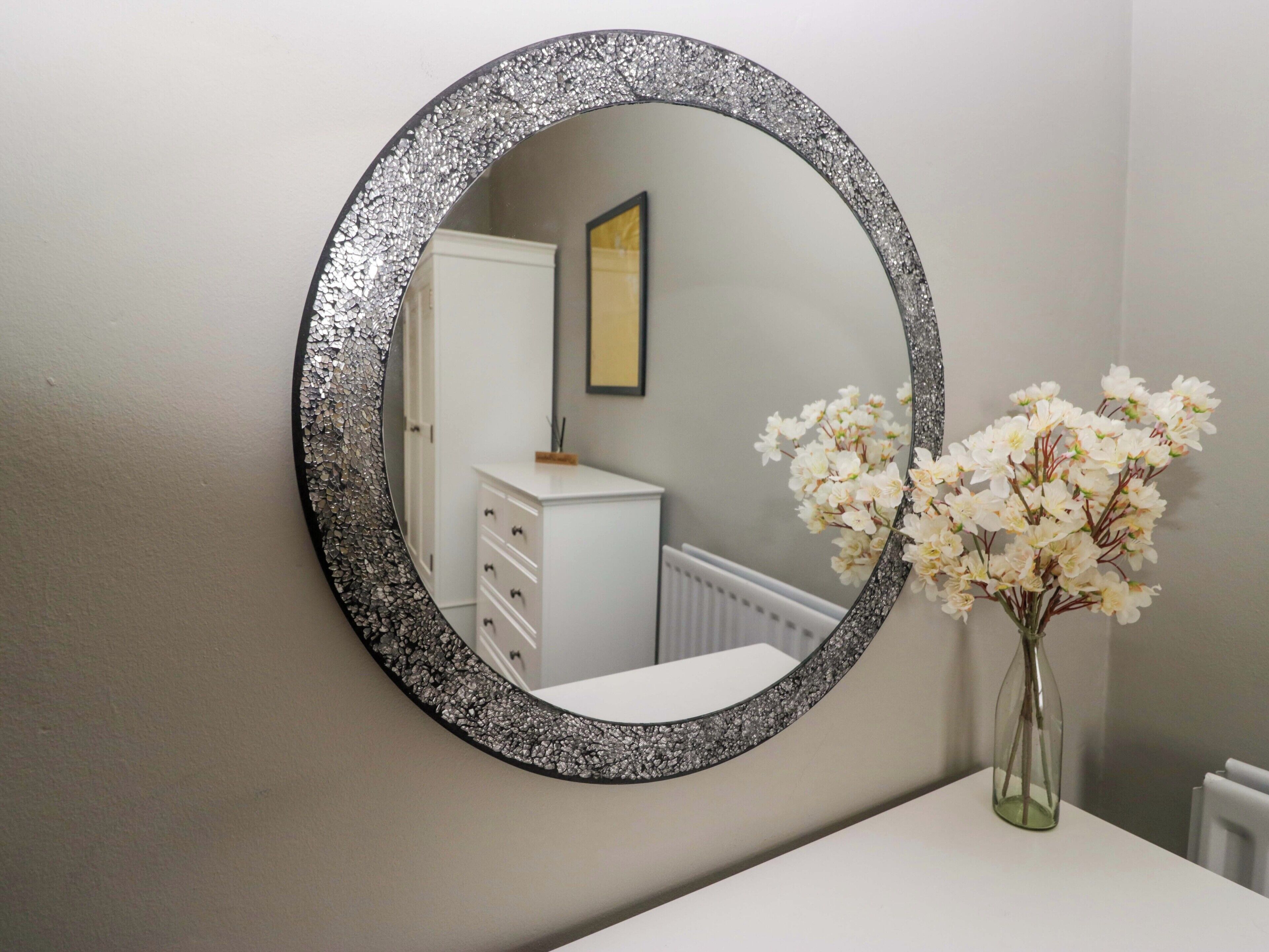 Un miroir rond avec un cadre texturé, une commode blanche, un vase avec des fleurs blanches et un tableau encadré accroché au mur.