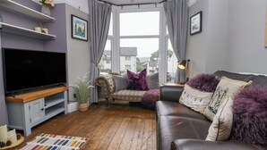 A living room with a TV, sofa, and a view of houses through large windows. - Poppy Cottage (Windermere)