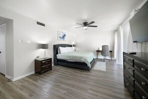 A bedroom with a bed, a desk, a chair, a dresser, a ceiling fan, and a painting on the wall.