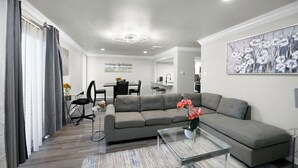 Una sala de estar moderna con un sofá gris, una mesa de centro de vidrio y un área de comedor al fondo.