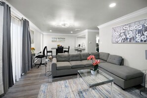 A modern living room with a grey sofa, a glass coffee table, and a dining area in the background.
