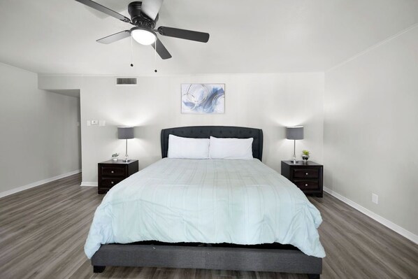 Un dormitorio moderno con una cama, dos mesitas de noche, un ventilador de techo y una obra de arte en la pared.