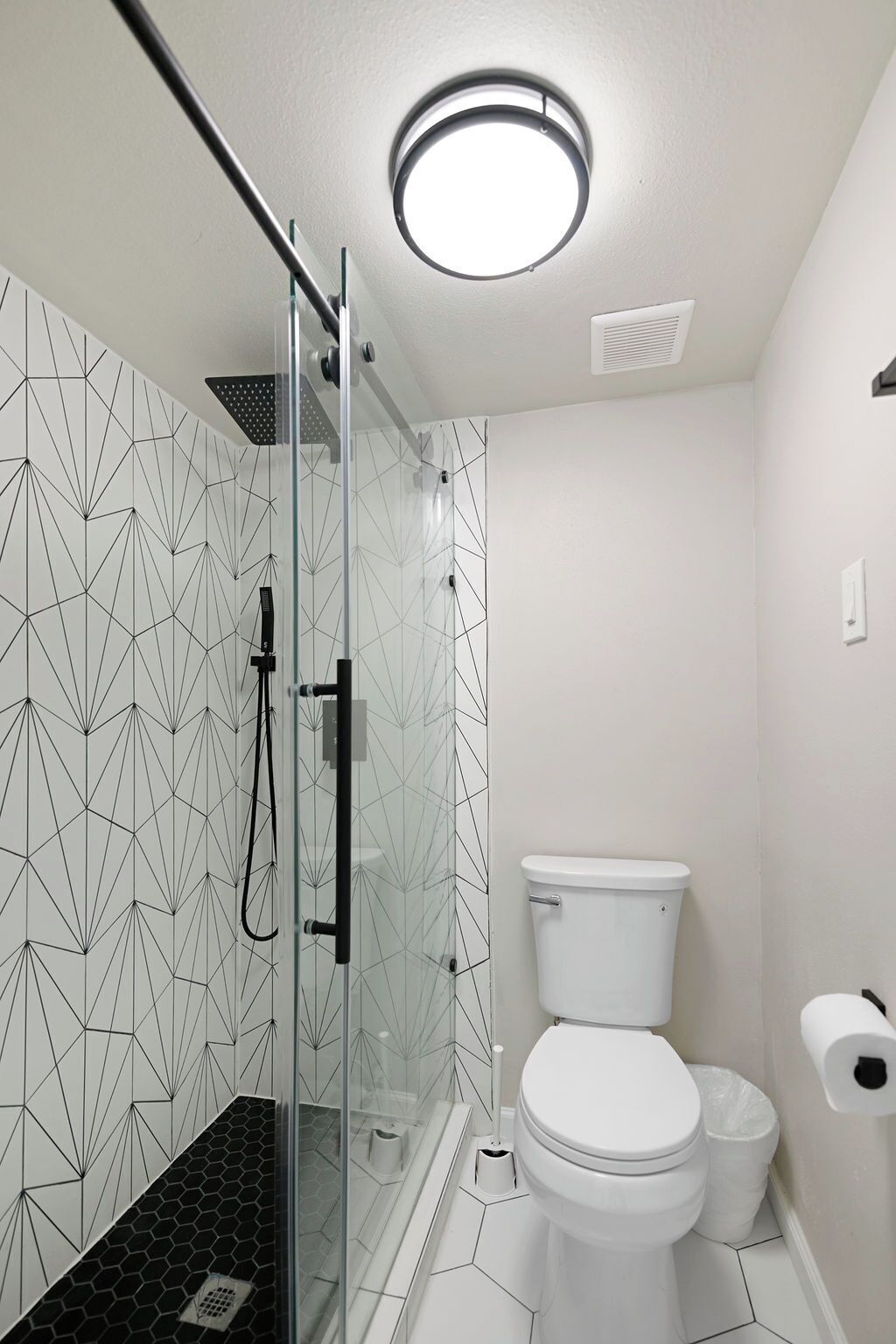 A modern bathroom with a geometric tile backsplash, a glass shower enclosure, a white toilet, and a toilet paper holder.