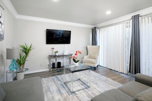 Una sala de estar moderna con televisor de pantalla plana, una mesa de centro de vidrio, un sillón color beige y un ventanal con persianas verticales.