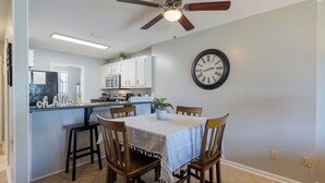 Un comedor con una mesa preparada para cuatro personas, un ventilador de techo y una cocina al fondo.