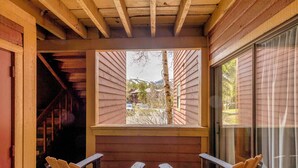 Un porche de madera con dos sillas y un banco, una ventana que ofrece vistas a una montaña y coches aparcados, y una escalera de madera a la izquierda.