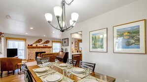 Un comedor con una mesa de madera dispuesta para cuatro personas, rodeada de sillas. Se observa una chimenea al fondo y dos cuadros en la pared.