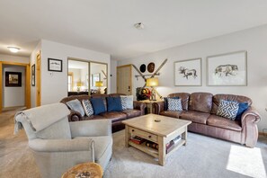 Ein Wohnzimmer mit einer braunen Ledersofa, einem hellen Sessel und einem Holztisch. An den Wänden befinden sich Bilder in Rahmen und ein Spiegel über dem Sofa.