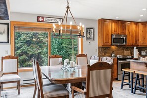 A dining area with a wooden table and chairs, a chandelier, and a view of trees through a large window.