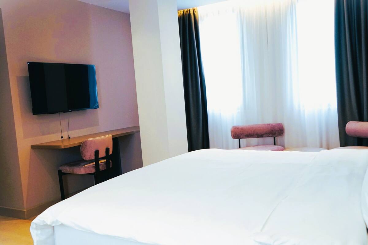 Une chambre d’hôtel avec un grand lit, un téléviseur à écran plat fixé au mur, un petit bureau avec une chaise et une fenêtre avec des rideaux.