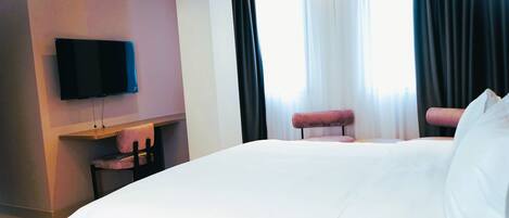 A hotel room with a large bed, a flat-screen TV mounted on the wall, a small desk with a chair, and a window with curtains.