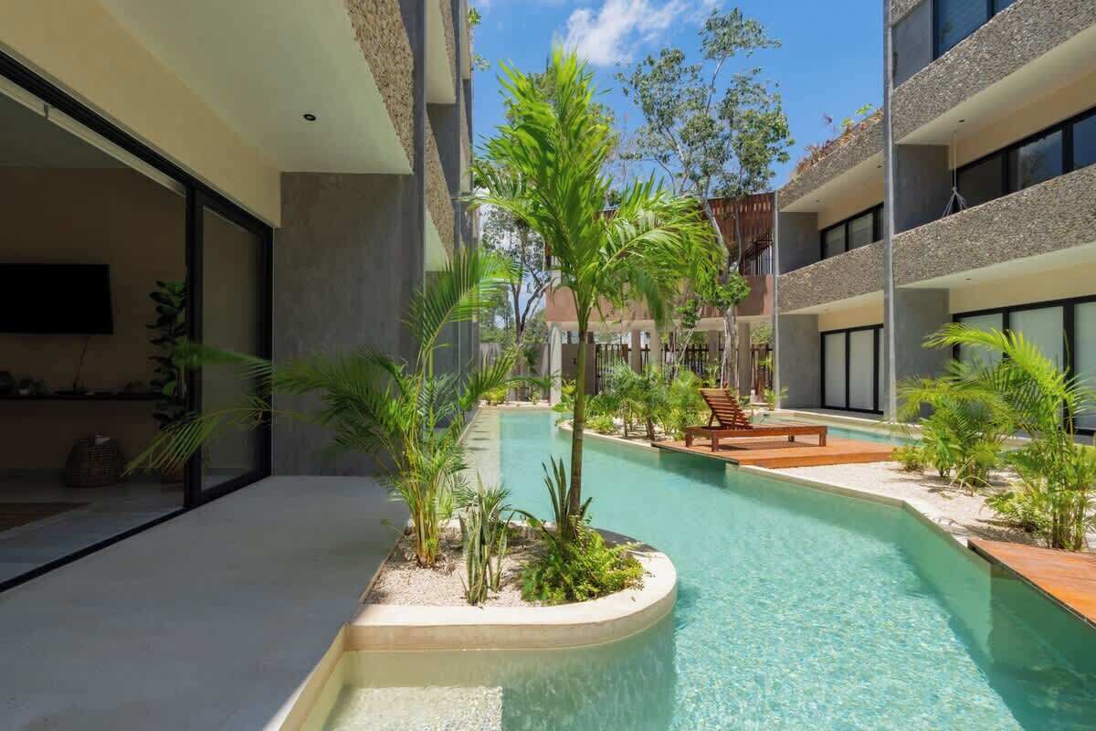 Une cour d’hôtel moderne dotée d’une piscine, entourée de palmiers et de plantes, et surmontée d’une structure à toit plat.