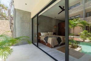 A modern bedroom with a large bed, a ceiling fan, and a view of a swimming pool and greenery.