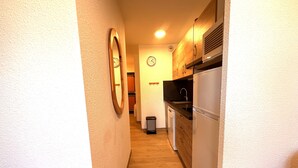 A narrow hallway leading to a kitchen with wooden cabinets, a refrigerator, and a clock on the wall.