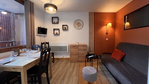 Un salon chaleureux avec un canapé, une table à manger et des chaises, un petit tabouret, un téléviseur fixé au mur et une horloge décorative.