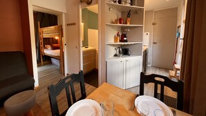 Une pièce compacte comprenant un lit superposé, une table à manger avec des chaises, un coin cuisine avec des étagères et un petit espace salon.