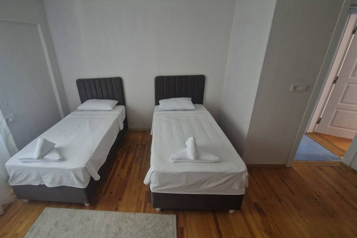 A room with two single beds, each with a pillow and a folded towel. The room has wooden flooring and a white rug.