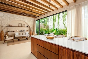 A modern kitchen with a large island, stone wall, wooden beams, and a view of greenery.