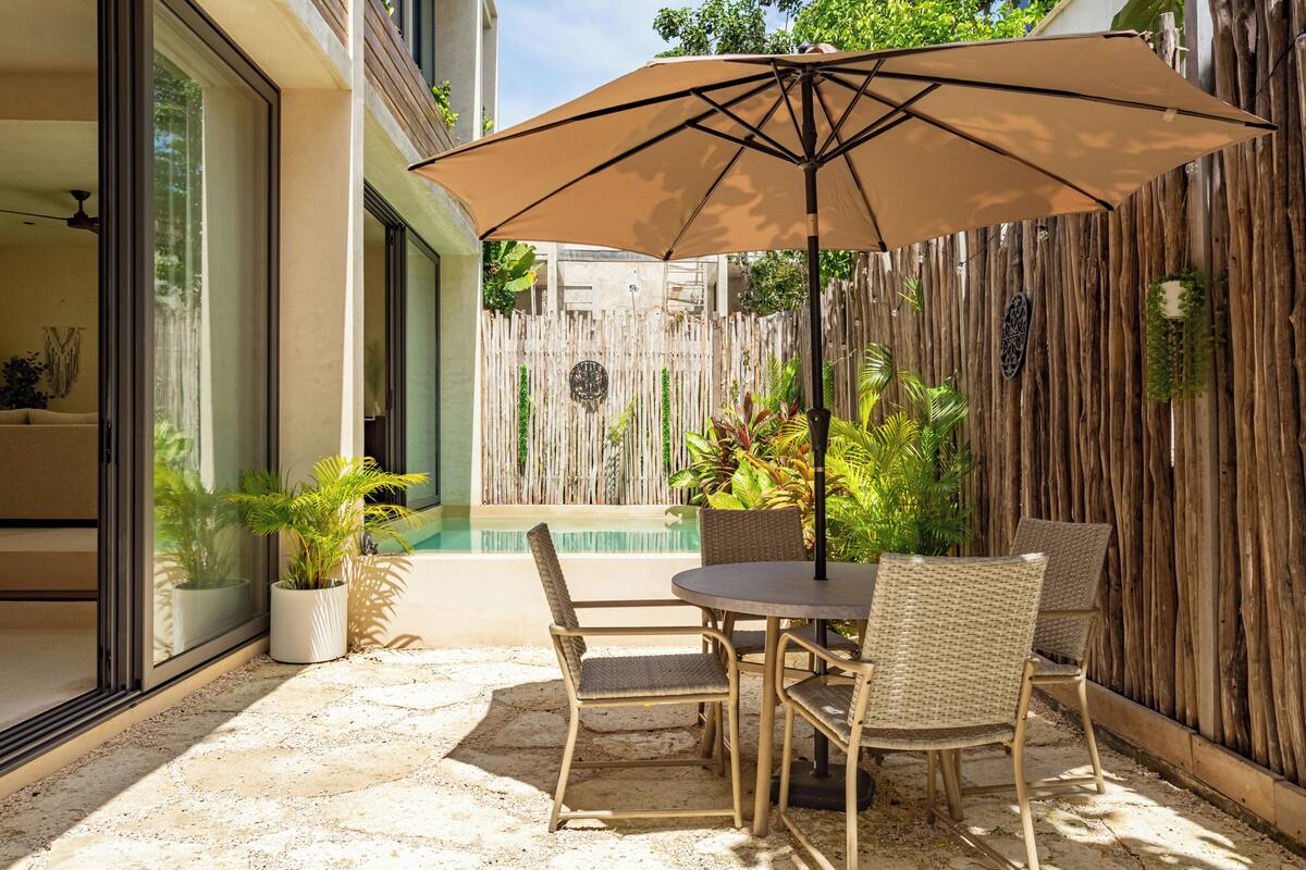 An outdoor patio with a table and chairs, a large umbrella, and a pool.