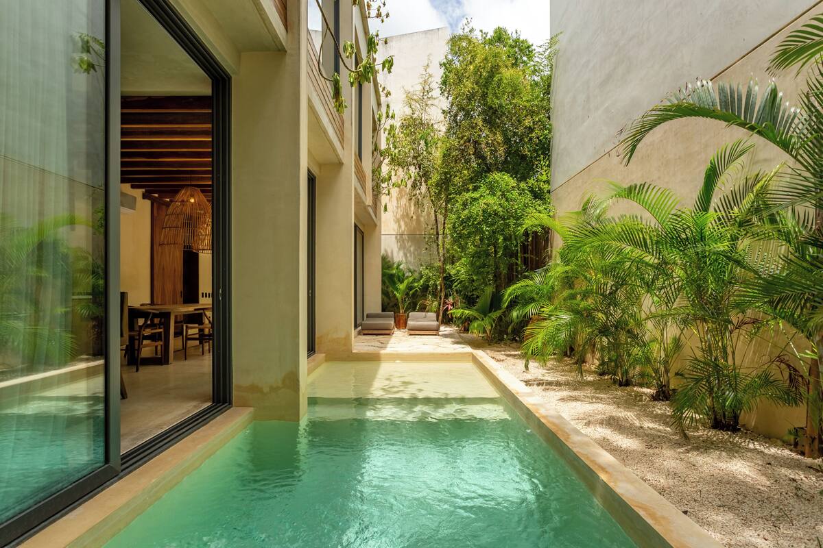 A narrow pool with a glass wall on one side and a building on the other, surrounded by greenery.