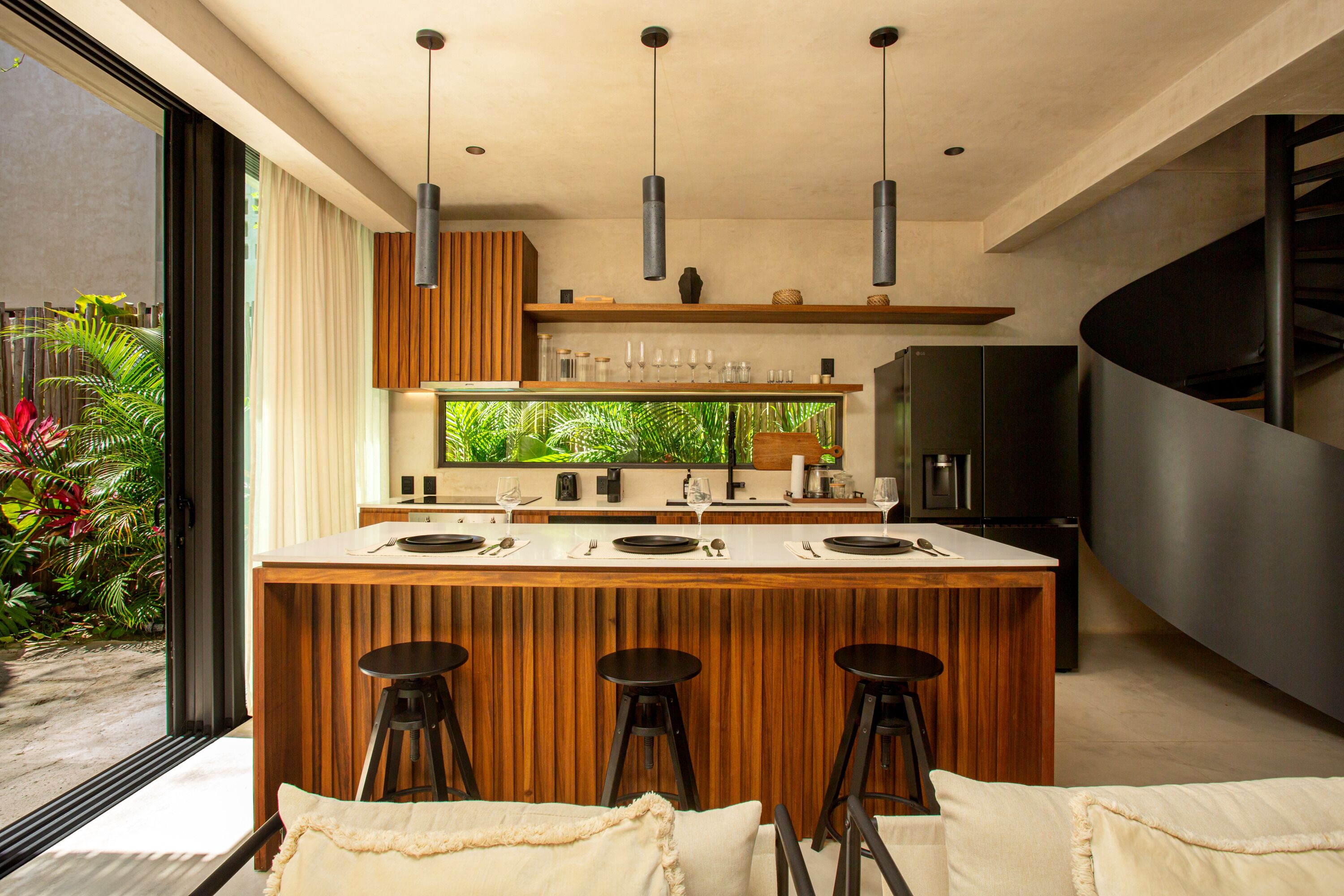 A modern kitchen with a central island, bar stools, pendant lighting, and a view of greenery outside.