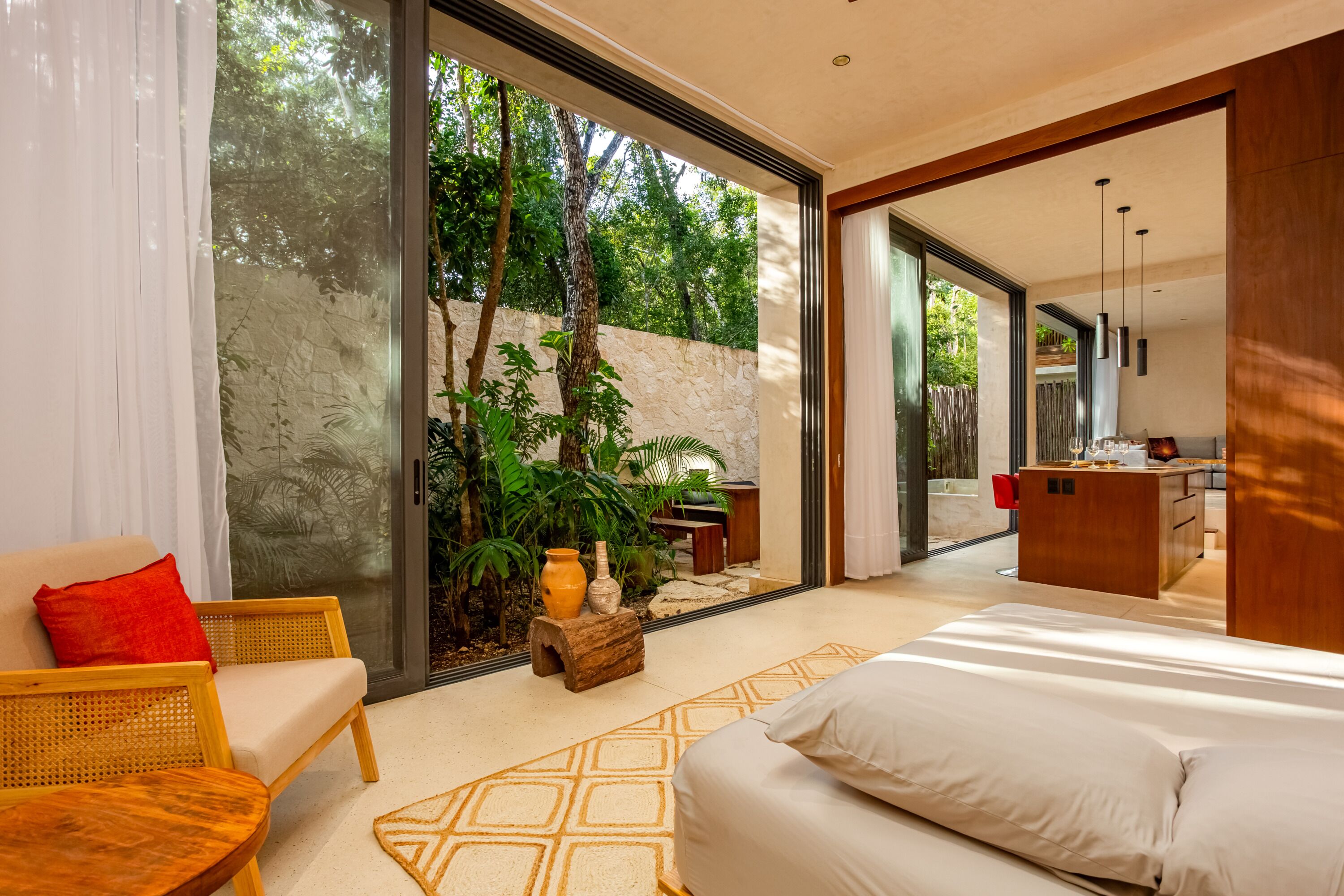 A modern bedroom with a large bed, a sitting area with a chair and table, and a view of a garden through a glass door.