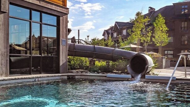 Una piscina con un tobogán de agua, un edificio de madera con grandes ventanales y un edificio de ladrillo al fondo.