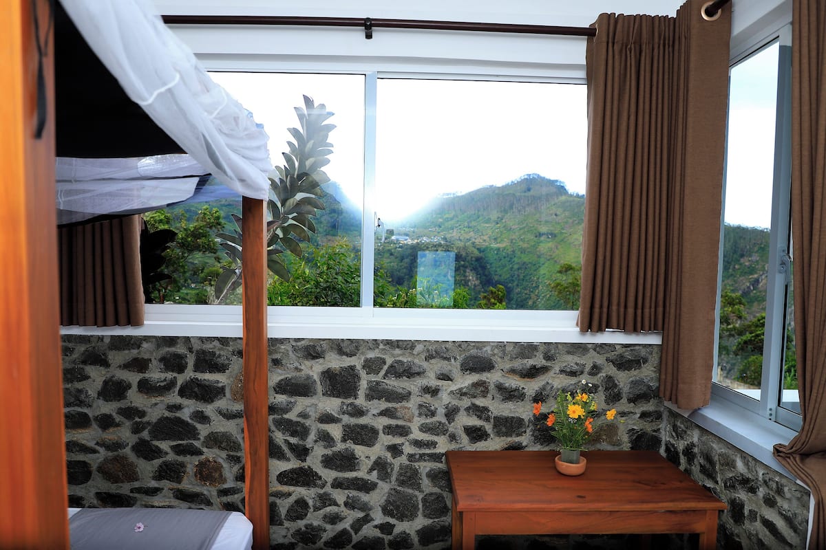 Una habitación con literas, una pared de piedra, una ventana con cortinas y una pequeña mesa con un jarrón de flores.
