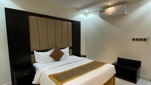 Una habitación de hotel moderna con una cama grande, cabecera con listones verticales, mesita de noche, unidad de aire acondicionado y un pequeño mueble de almacenamiento negro.