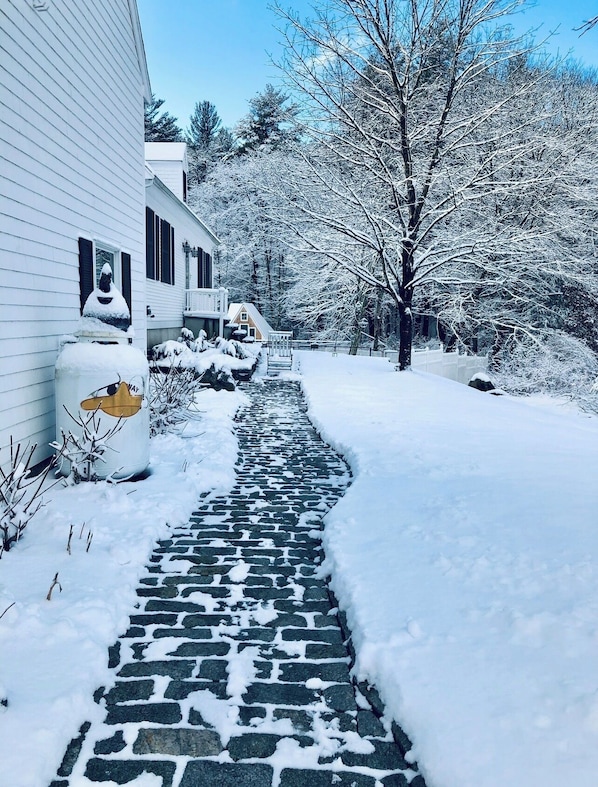 A snow-covered pathway leading to a white house with a chimney, surrounded by trees and a small bridge.