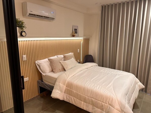 A neatly made bed with a beige comforter and pillows, a bedside table with a clock and a small plant, a wall-mounted air conditioner, and a window with vertical blinds.