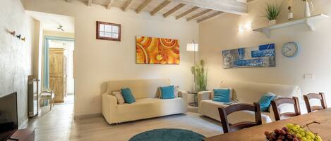 A living room with a wooden ceiling, a beige sofa, a dining table with a bowl of fruit, and a blue rug.