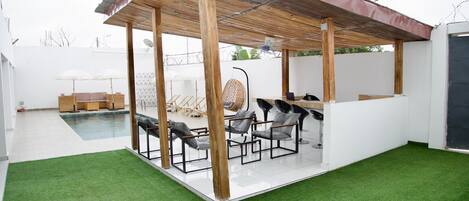 A covered outdoor seating area with a pool, artificial grass, and a white wall in the background.