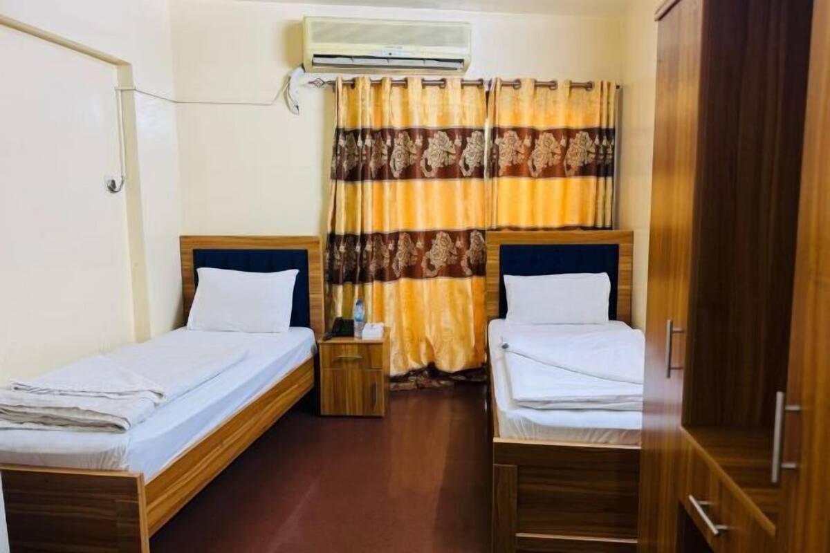 A room with two single beds, a wooden wardrobe, a nightstand, and an air conditioning unit.